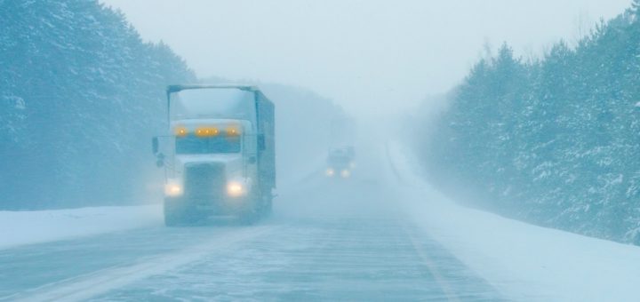 A semi-truck with its headlights on, driving down an icy road. It's snowing heavily, and visibility is low.