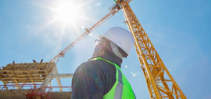 A construction worker wearing a safety vest and a hard hat outside. The worker is looking up at a yellow crane.