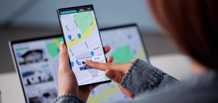 A smartphone displaying a real estate map with blue pins and $1.1M listing; a laptop in the background shows a similar view.
