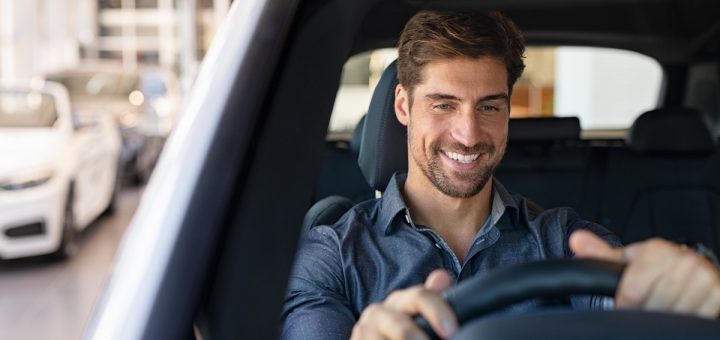 A man wearing a blue polo shirt smiles as he test-drives a car in a dealership. Behind are rows of other cars.