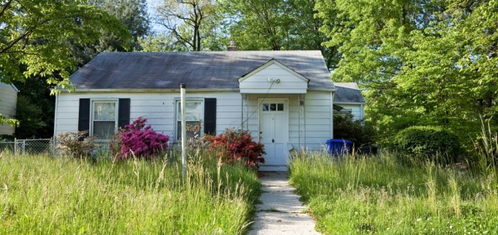 A small, dilapidated house with a small walkway. The grass is highly overgrown and creeping onto the walkway.
