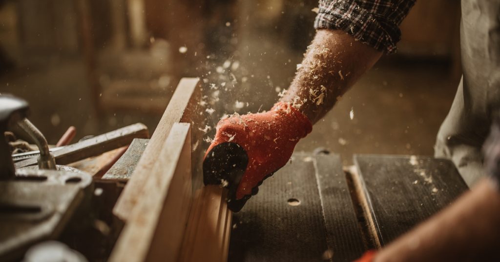 A carpenter's hands push a long piece of wood through a saw blade. The carpenter's arm is covered in wood shavings.