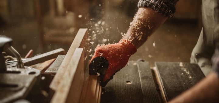 A carpenter's hands push a long piece of wood through a saw blade. The carpenter's arm is covered in wood shavings.