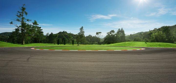 A ground-level view of a blacktop circle racetrack with green grass and trees surrounding it on a sunny day.