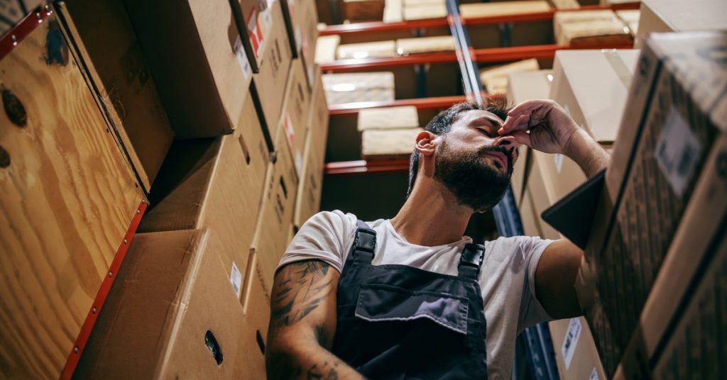 A man is sitting next to racks filled with cardboard boxes. His arm is resting on a table and he is pinching his nose.