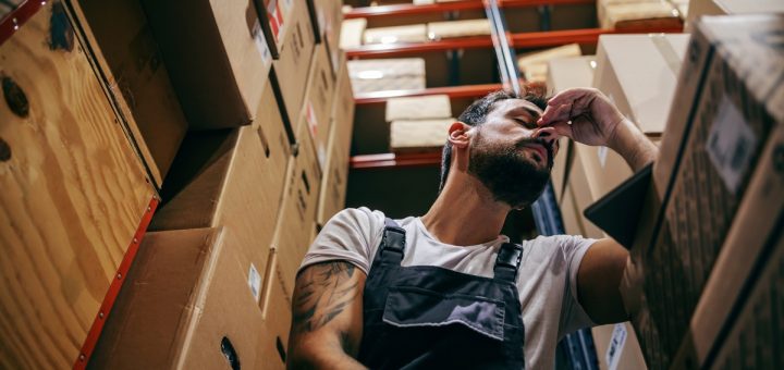 A man is sitting next to racks filled with cardboard boxes. His arm is resting on a table and he is pinching his nose.