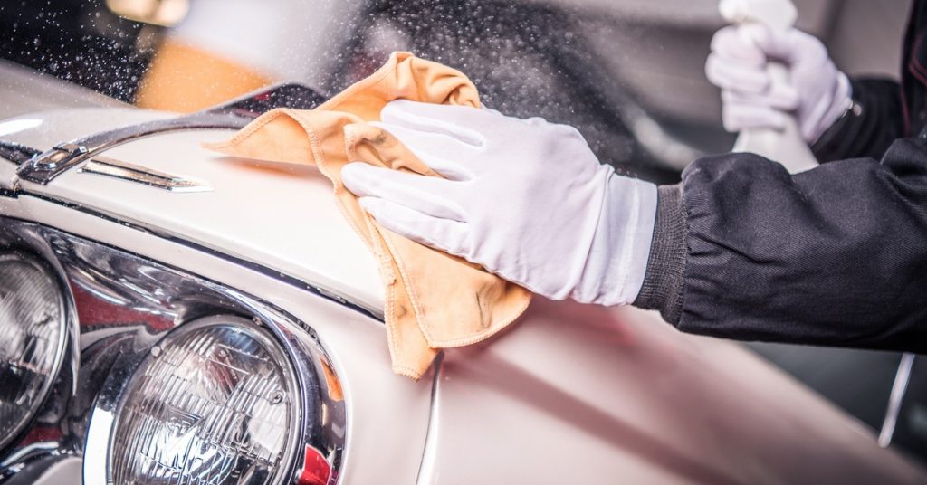 A gloved hand is holding a cloth while rubbing it against the body of a car. There is another hand holding a spray bottle.