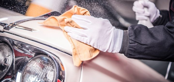 A gloved hand is holding a cloth while rubbing it against the body of a car. There is another hand holding a spray bottle.
