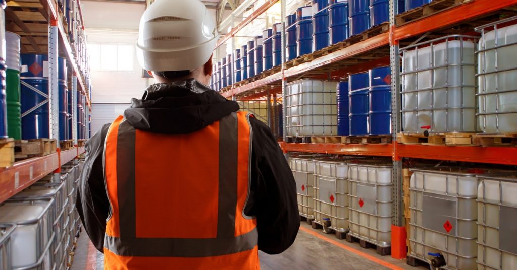 A man in a white hard hat and an orange vest is looking at racks filled with containers and barrels.