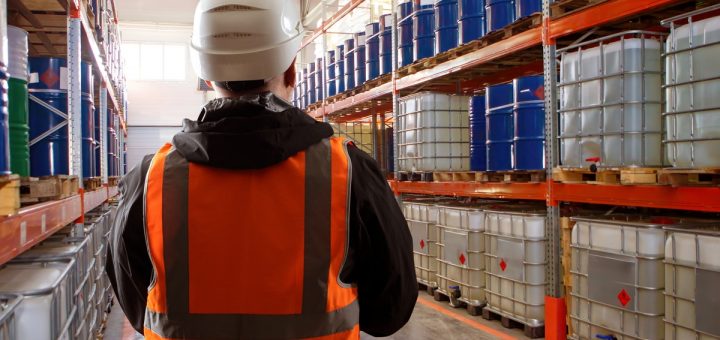 A man in a white hard hat and an orange vest is looking at racks filled with containers and barrels.