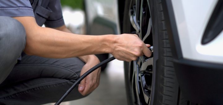 A man in a blue short-sleeved shirt and jeans crouching down to inflate a tire on his white vehicle.