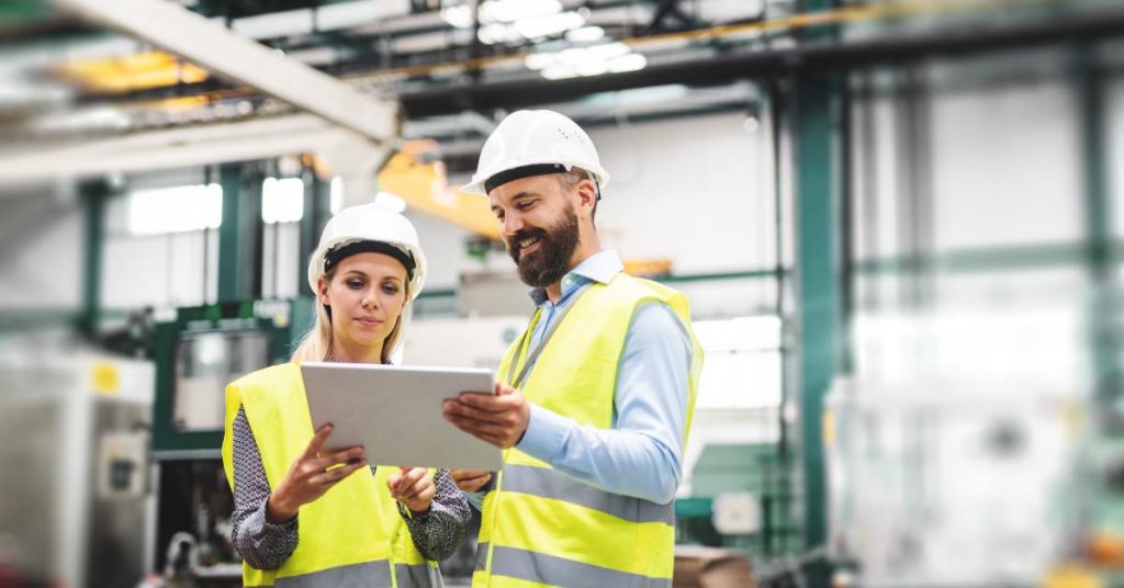 A man and a woman are standing next to each other; they are both wearing hard hats and high-visibility vests.