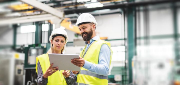 A man and a woman are standing next to each other; they are both wearing hard hats and high-visibility vests.
