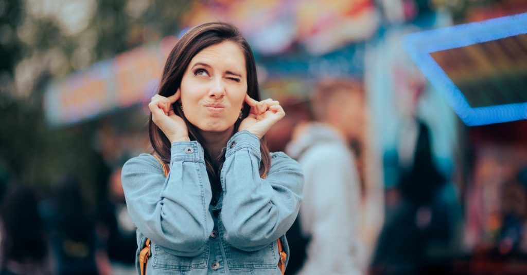 A woman wearing a denim shirt stands outside and plugs her ears with her fingers. The background is blurred.