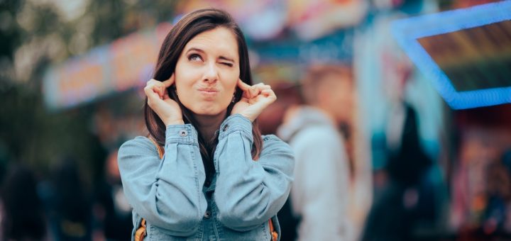 A woman wearing a denim shirt stands outside and plugs her ears with her fingers. The background is blurred.