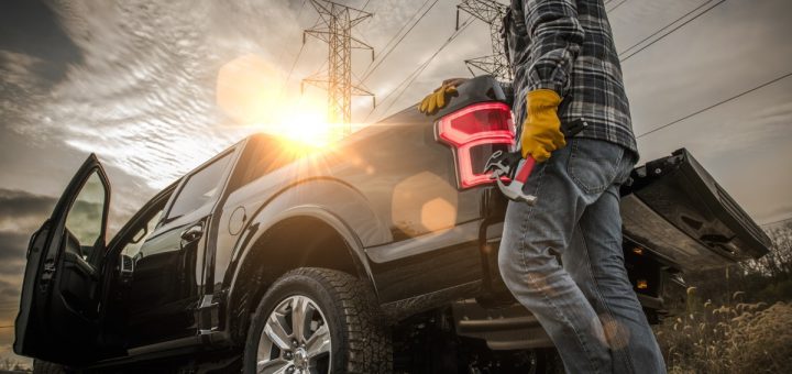 A low-angle view of a man wearing a hard hat gripping the bed of a black pickup truck parked near transmission towers.