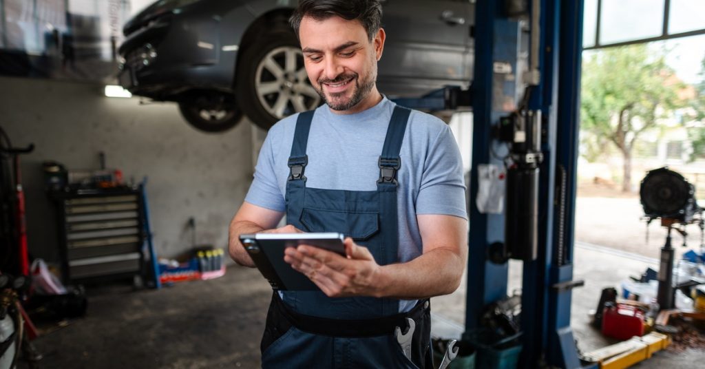 A mechanic smiling at tablet in their hands is standing in the middle of an auto repair shop during the daytime.