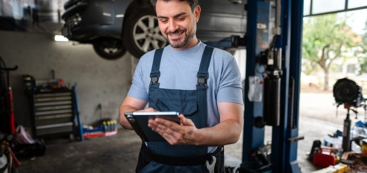 A mechanic smiling at tablet in their hands is standing in the middle of an auto repair shop during the daytime.