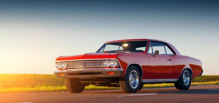 A red classic muscle car with a chrome grille parked on an asphalt road next to an open grassy field.