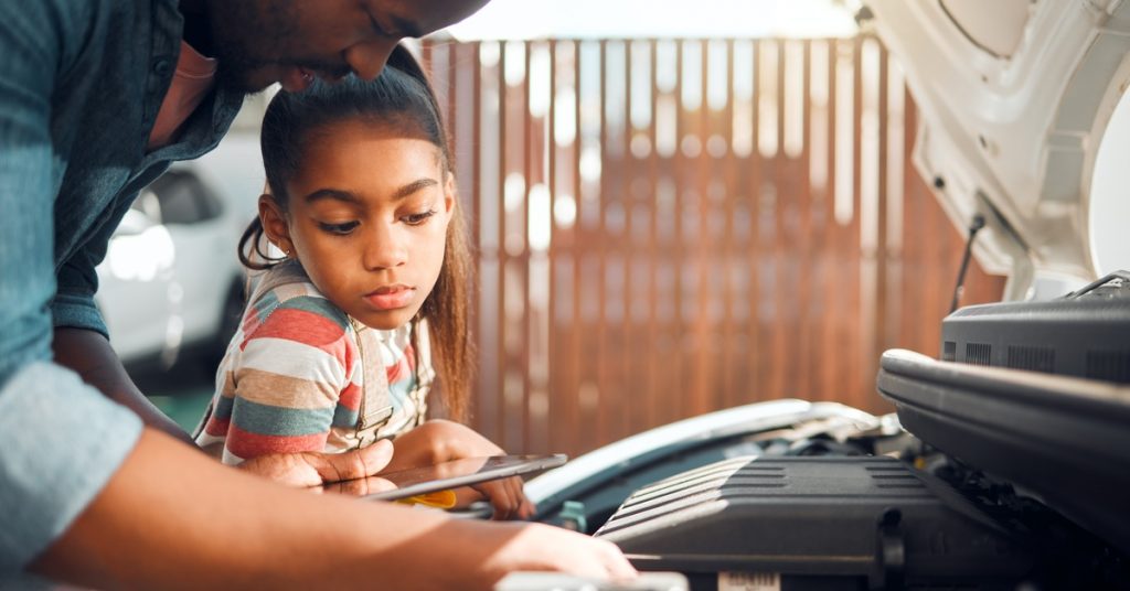 A father and daughter looking at the internal engine of a car. The hood of the car is up above them.