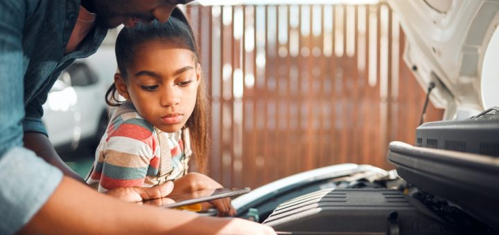 A father and daughter looking at the internal engine of a car. The hood of the car is up above them.