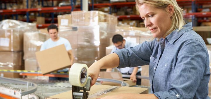 A woman in the foreground is taping up a cardboard box. There are others in the background who are doing the same.
