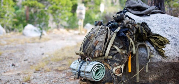 A fully packed camouflage-patterned hiking backpack leaned up against a large boulder off to the side of a trail.