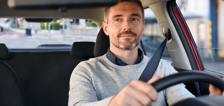 A man driving a car with both hands on the steering wheel, wearing a seatbelt and gray sweater, seen from inside the vehicle.