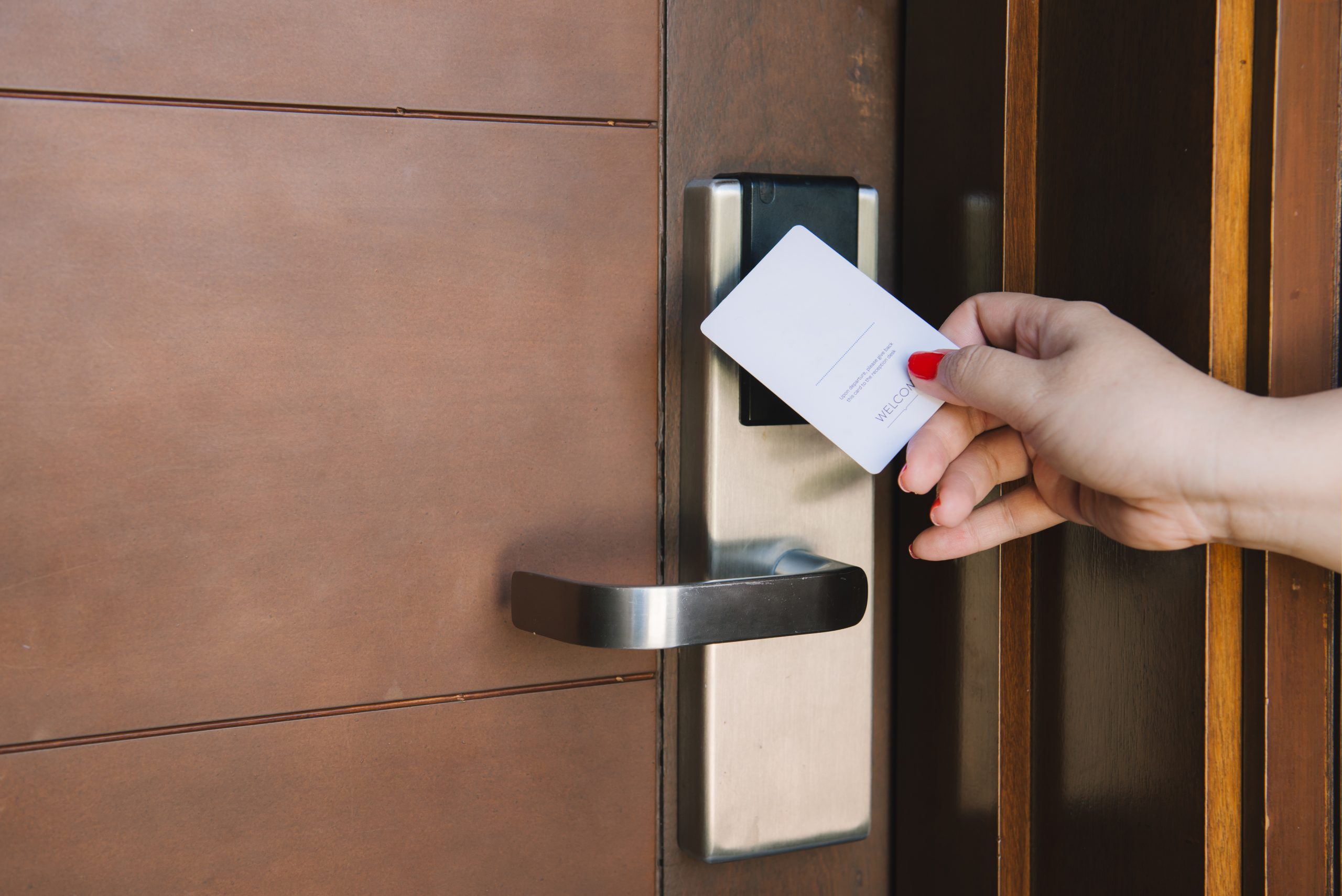 An individual holding a white keycard taps an electronic hotel door lock on a wooden door with a metal handle.