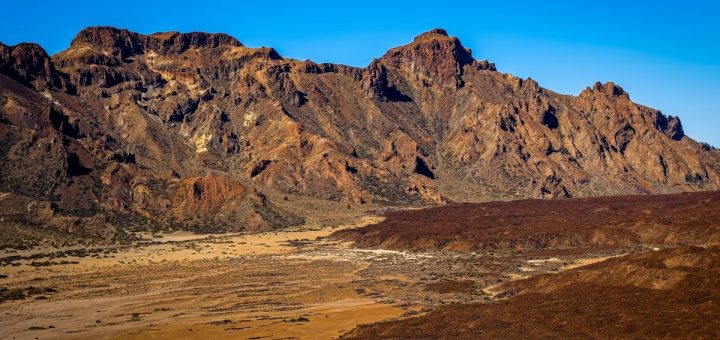 Rocky desert landscape with rugged brown mountains, dry sandy ground, and clear blue sky stretching overhead.