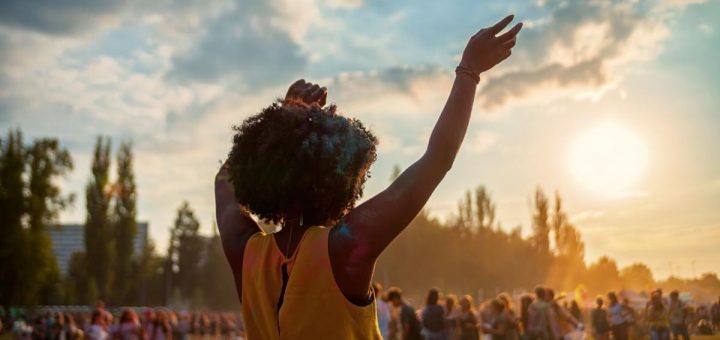 Back view of person dancing with arms raised at outdoor festival crowd during sunset under partly cloudy sky.