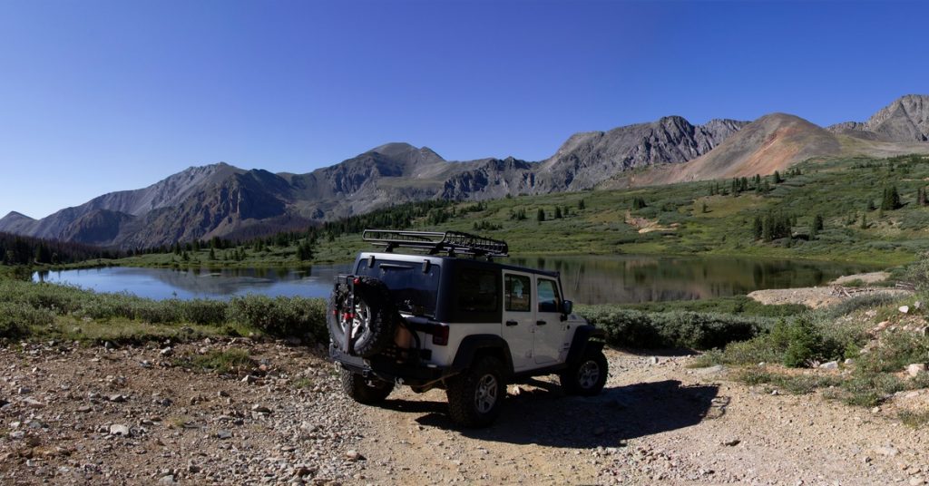 A rear view of a white Jeep with a black top parked near a lake on a sunny day with mountains in the background.