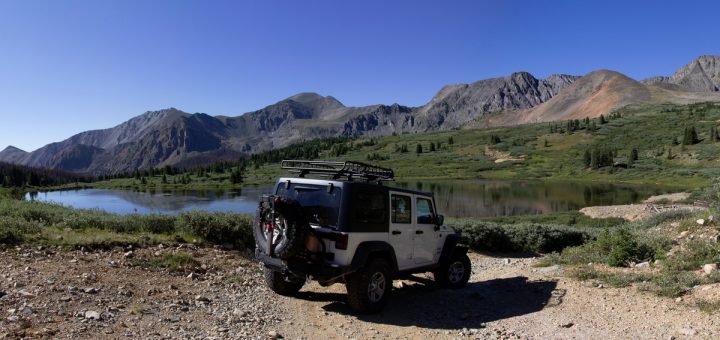 A rear view of a white Jeep with a black top parked near a lake on a sunny day with mountains in the background.