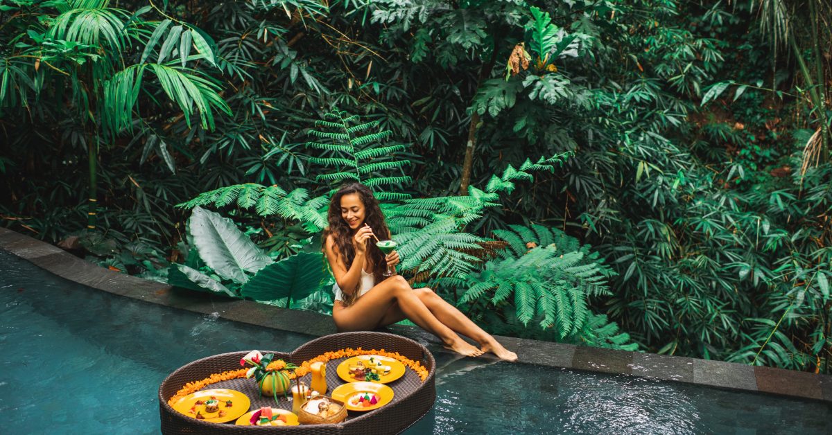 Woman relaxing by pool in jungle setting with drink, next to floating tray filled with colorful breakfast plates.