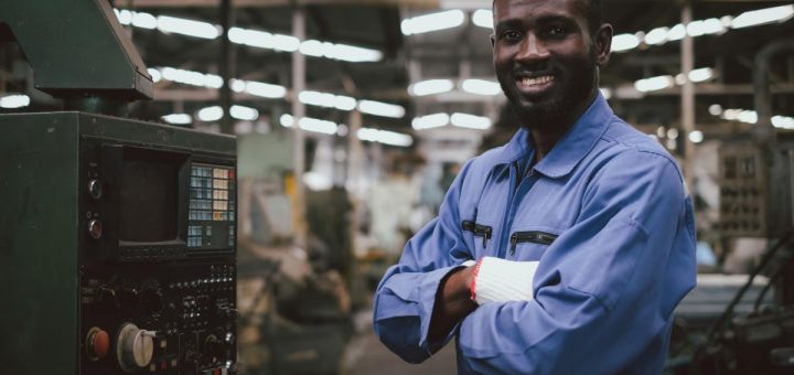A man in a blue jumpsuit and white gloves smiles and stands in front of a machine's controls inside a factory.