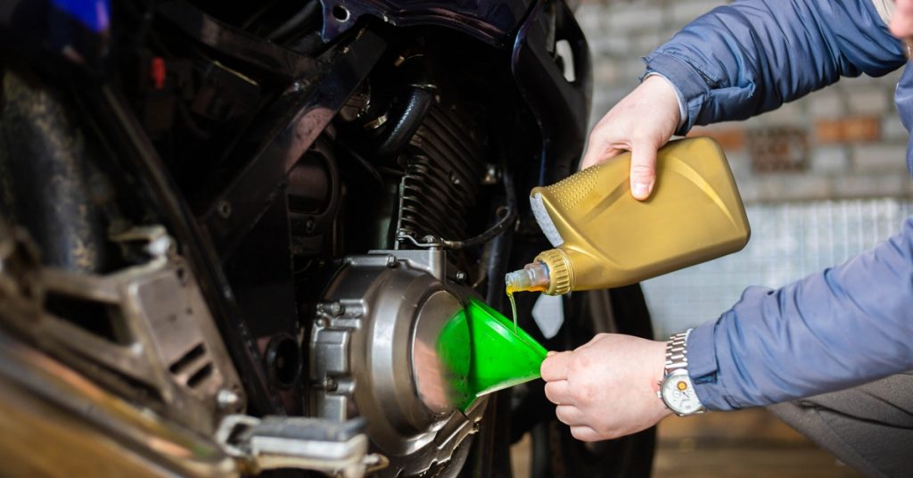 A close-up of a person pouring motor oil into the engine of a black motorcycle using a green funnel in a garage.