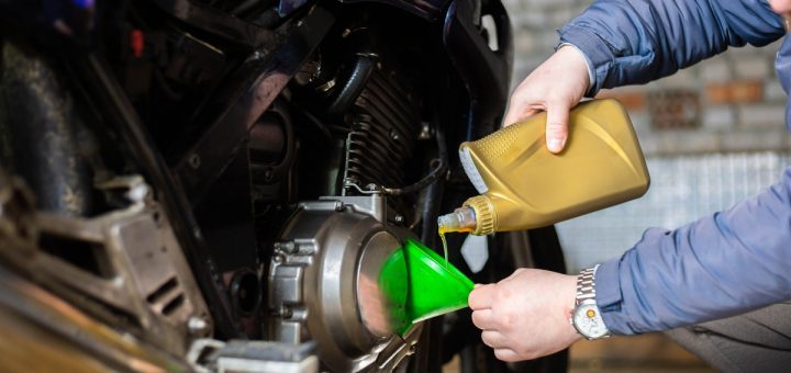 A close-up of a person pouring motor oil into the engine of a black motorcycle using a green funnel in a garage.