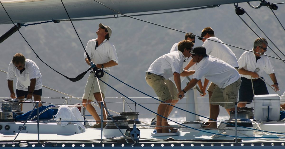 Seven men wearing white polo shirts and cargo shorts on top of a small yacht, tying ropes around the cover.
