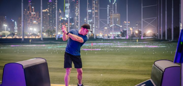 A man swinging a golf club at a lit driving range during the night with a city skyline and glowing targets on the field.