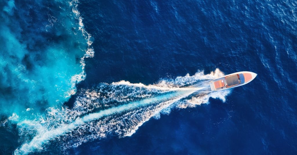 A white motorboat with a few people on it cruising across dark blue water, leaving a long white wake trailing behind it.