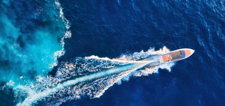 A white motorboat with a few people on it cruising across dark blue water, leaving a long white wake trailing behind it.