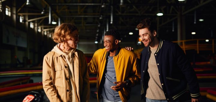 Three young men walk away from a large go-kart venue, smiling as the expansive track stretches behind them.