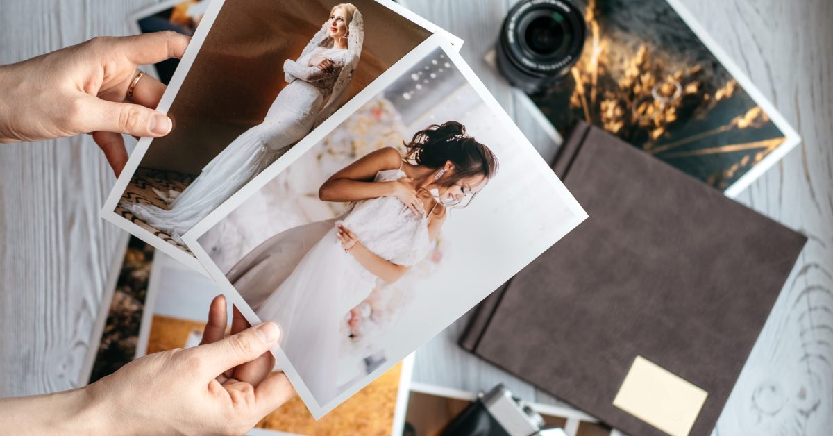 A person holds wedding photo prints, with several more scattered on the floor alongside a camera and an album.