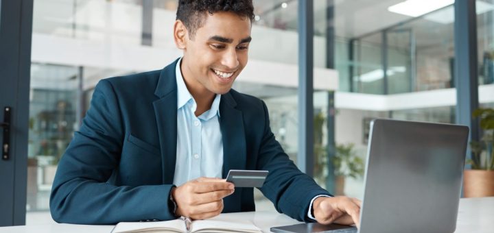 A person smiling and typing on a laptop keyboard with one hand. The person has a credit card in their other hand.