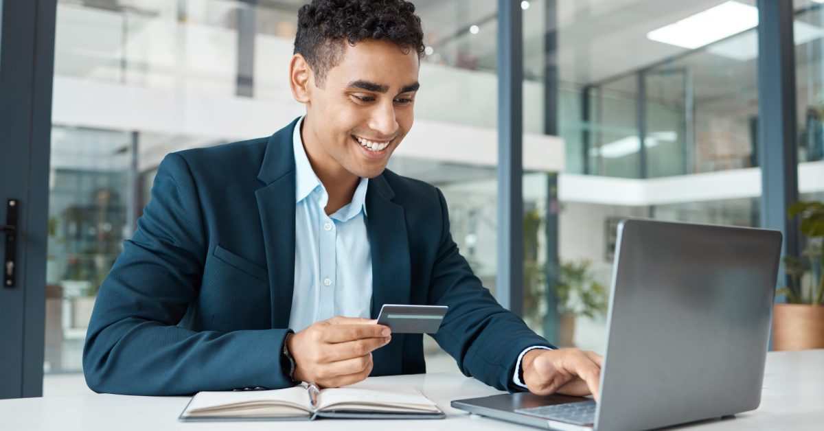 A person smiling and typing on a laptop keyboard with one hand. The person has a credit card in their other hand.