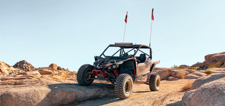 A dirt-covered side-by-side UTV on an off-road trail. One of its wheels is elevated onto a nearby rock.