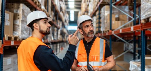 Two warehouse workers in hard hats and orange safety vests talking to one another inside of a warehouse.