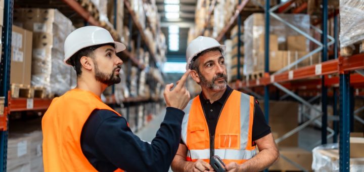 Two warehouse workers in hard hats and orange safety vests talking to one another inside of a warehouse.