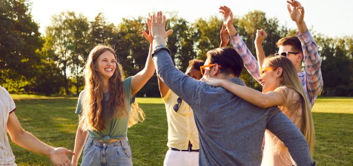Group of friends outdoors raise hands for high-five, smiling and celebrating together on sunny grassy field.