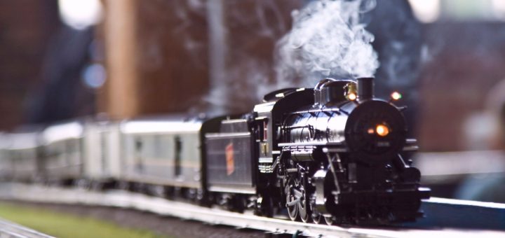 A closeup on a model steam train driving along a track. A plume of steam is rising from the train's smokestack.